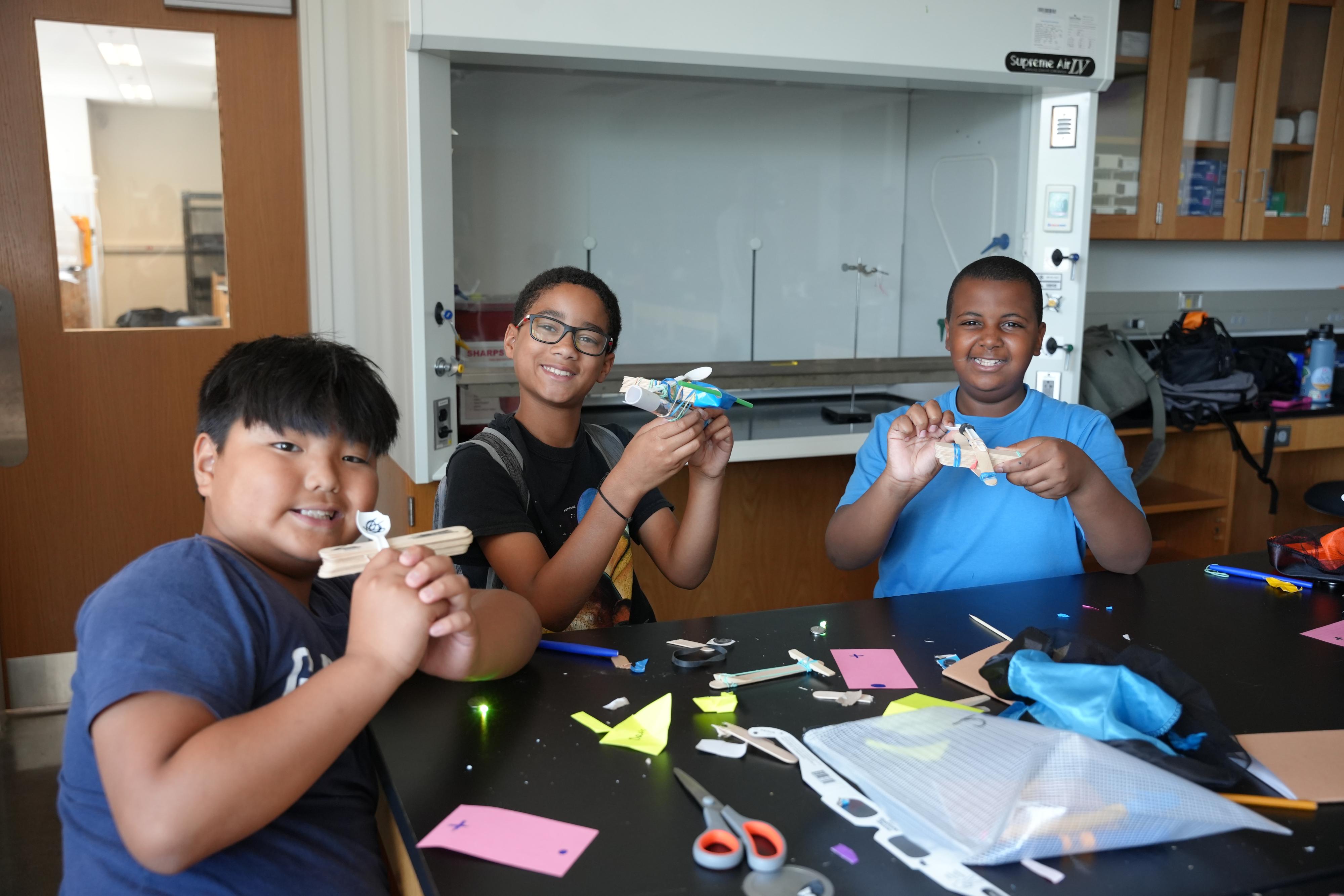 Students posing with their creations while sitting at a table.